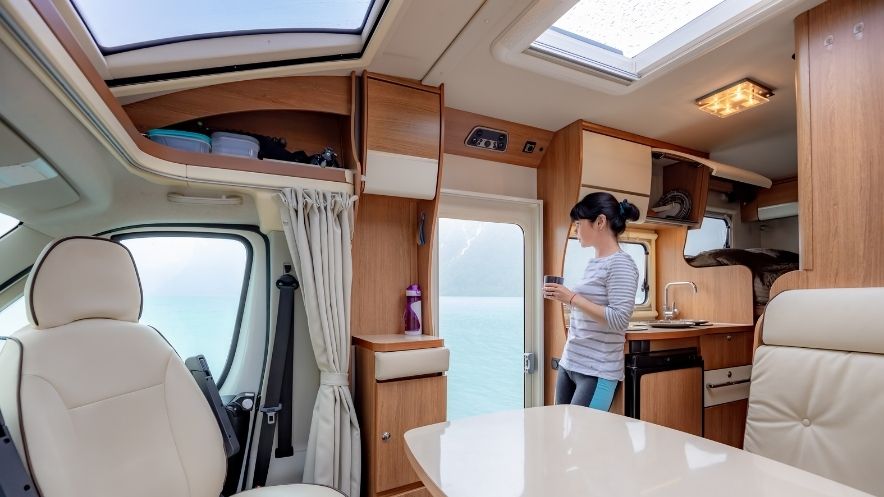 Young woman standing in an RV Camper with a drink looking out onto a lake. 