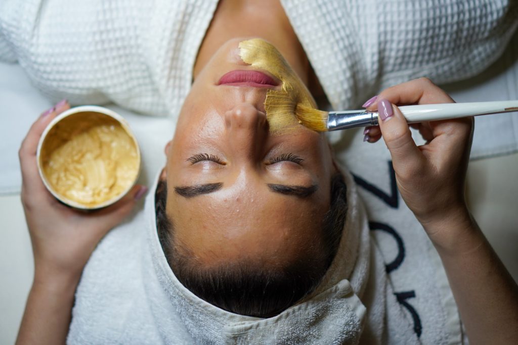 Woman laying with eyes closed receiving a gold mask treatment to her face.